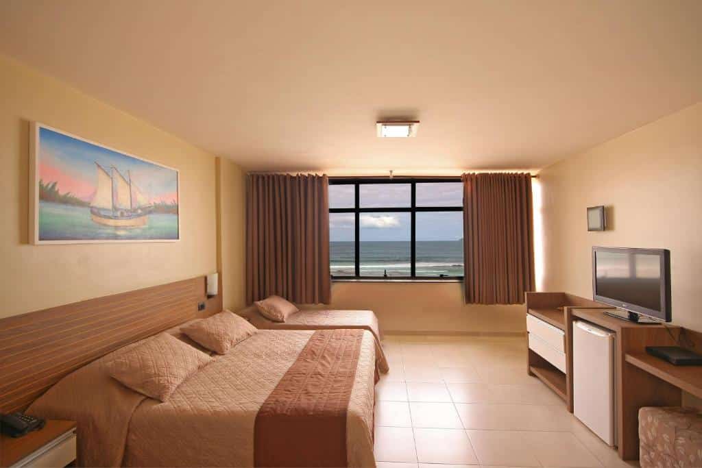 Quarto espaçoso no Malibu Palace Hotel com uma casa de casal, uma cama de solteiro, quadro decorativo, cômoda com frigobar, televisão e grande janela com cortinas e vista para o mar, para representar hotéis em Cabo Frio.