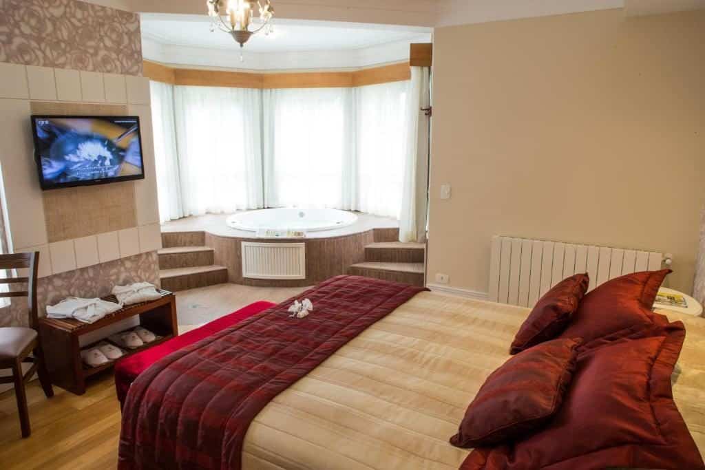 Um quarto no Carballo Hotel & Spa com uma cama grande, uma TV na parede, uma jacuzzi com janelas panorâmicas e uma cadeira com chinelos embaixo, para representar hotéis em Campos do Jordão.