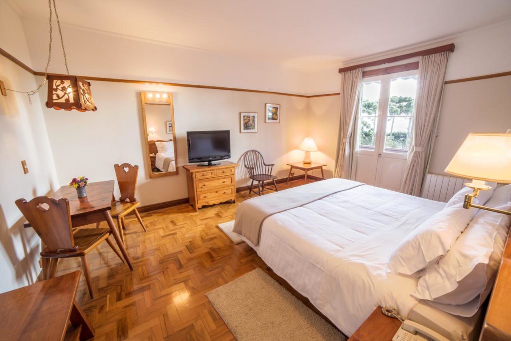 Quarto no Hotel Toriba amplo, com uma sacada lateral com cortinas, uma cama de casal, 7ma mesa com duas cadeiras de madeira do lado esquerdo, próximo há um espelho e um móvel de madeira com uma televisão.