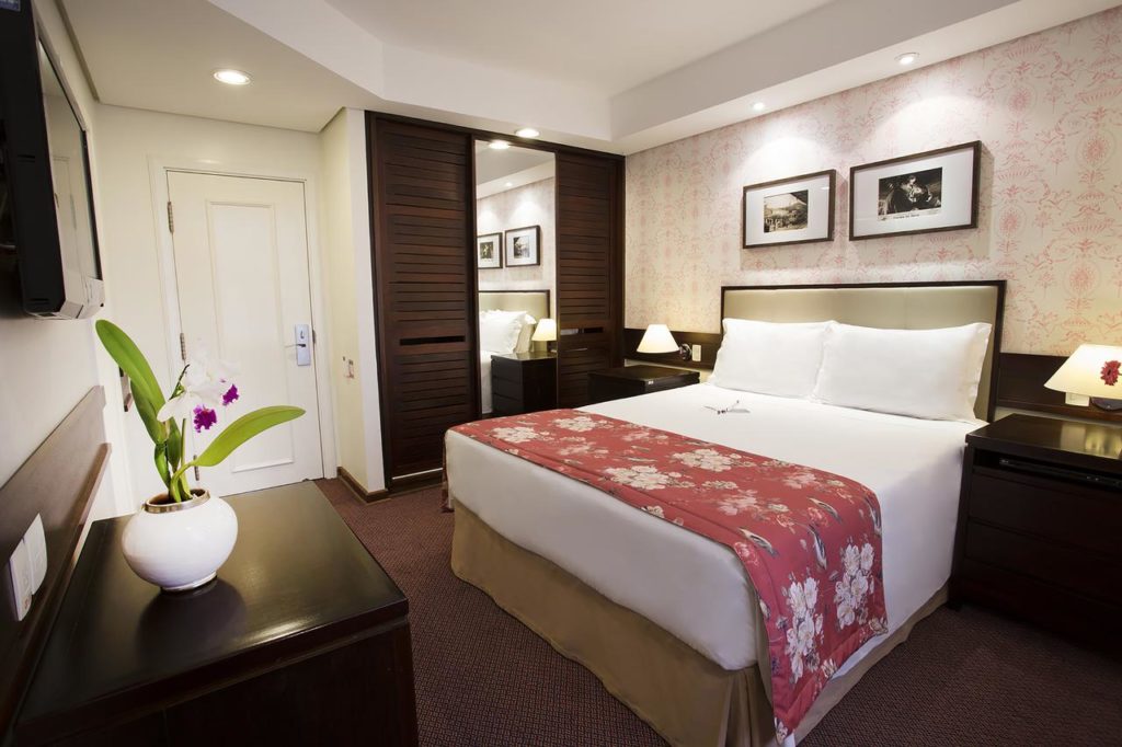 Quarto do Hotel Vila Inglesa com uma cama de casal moderna, um armário com espelho, duas mesinahs de cabeceira com abajures, além de um móvel de madeira de frente para a cama, onde há uma televisão presa na parede, para representar hotéis em Campos do Jordão.