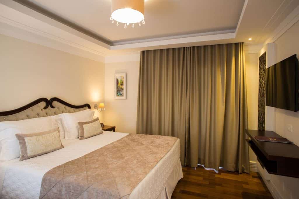 Quarto do Hotel Estoril com uma cama grande, roupa de cama bege, TV na parede, cortinas e uma luminária de teto.
