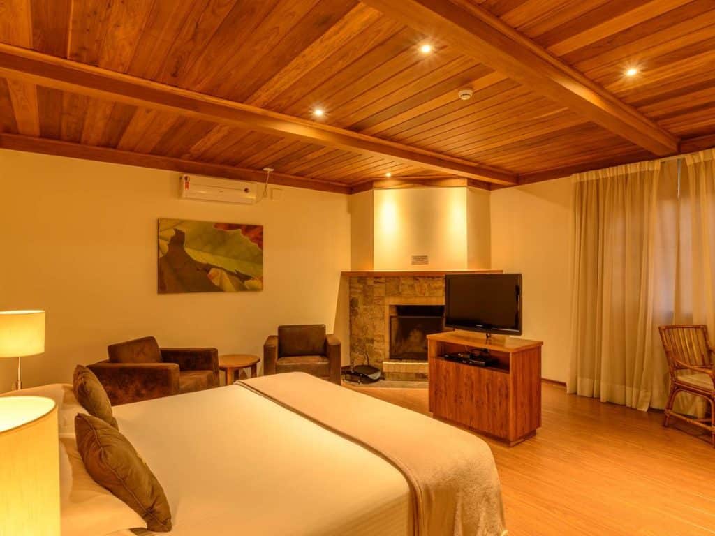Quarto no Le Renard com iluminação aconchegante, painéis de madeira, cama, poltronas, lareira, TV e cortinas grandes.