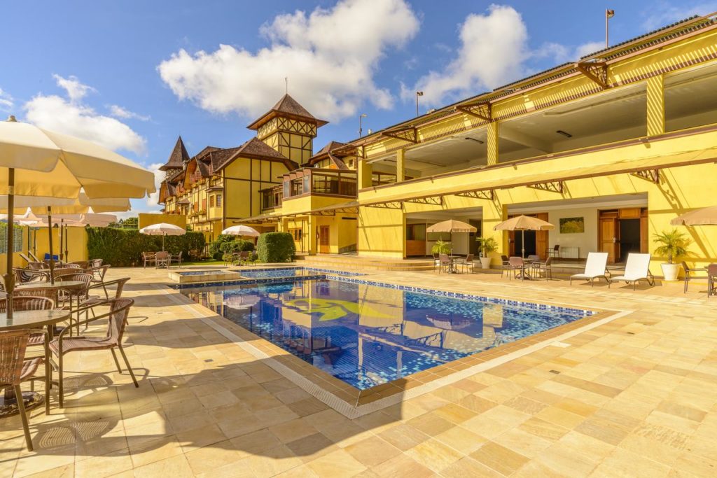 Piscina externa do Le Renard com espreguiçadeiras, mesas e guarda-sóis ao redor, ao lado de um grande edifício amarelo do hotel, sob um céu azul.