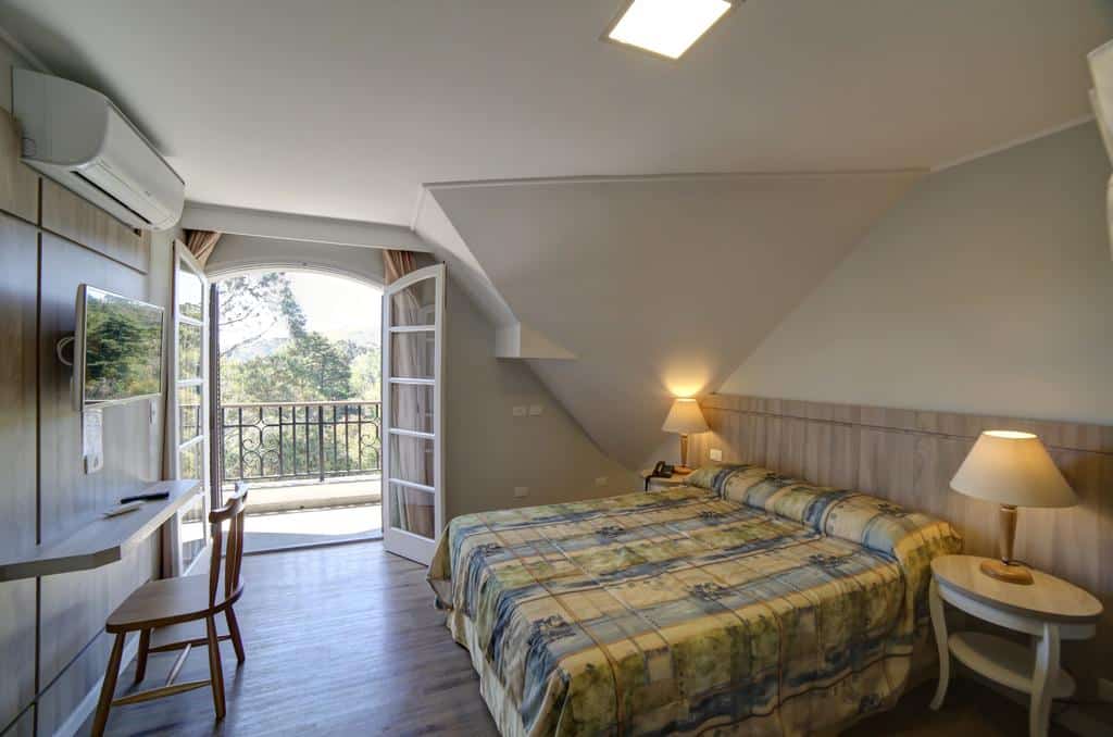 Quarto do Moinho Itália Hotel com uma cama, duas lâmpadas, uma TV, uma escrivaninha com uma cadeira e uma porta aberta que dá para uma varanda com vista.