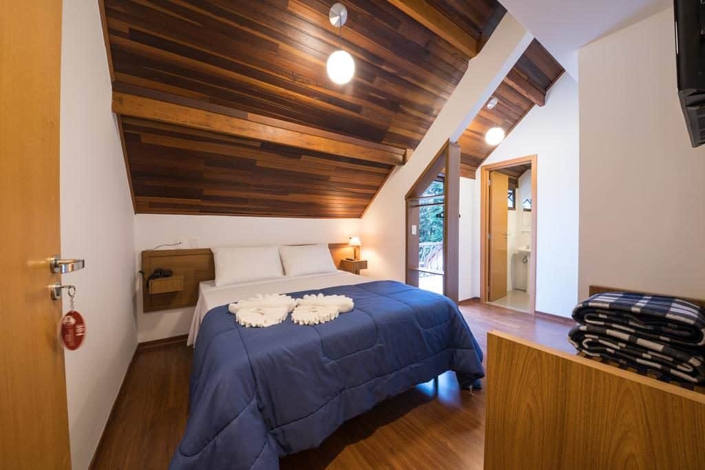 Quarto na Vila Dom Bosco com teto inclinado de madeira, cama de casal com edredom azul e toalhas dobradas sobre a cama.