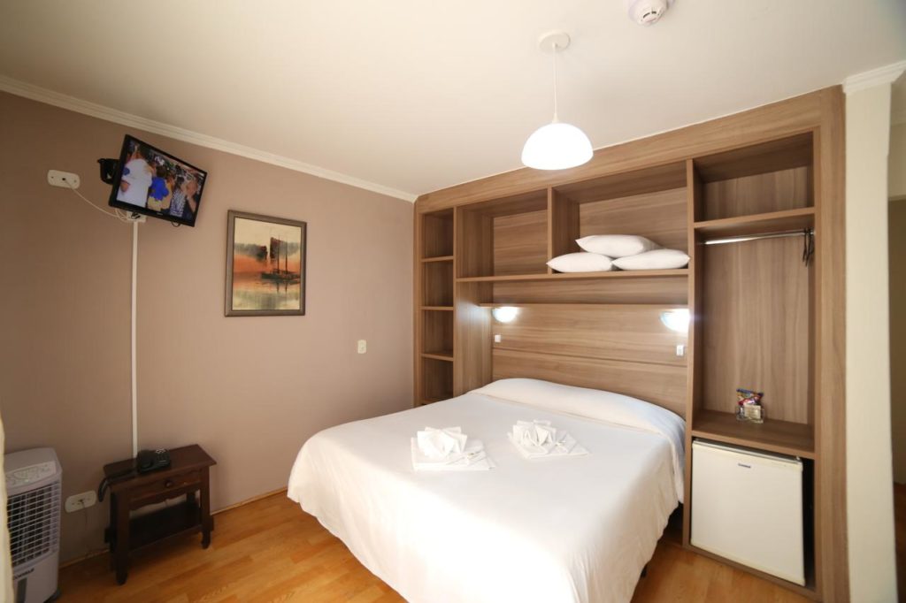 Quarto do Hotel JB com uma cama de casal, prateleiras nas paredes, um pequeno frigobar, uma TV, uma mesa de cabeceira e um quadro na parede, para representar hotéis em Campos do Jordão.