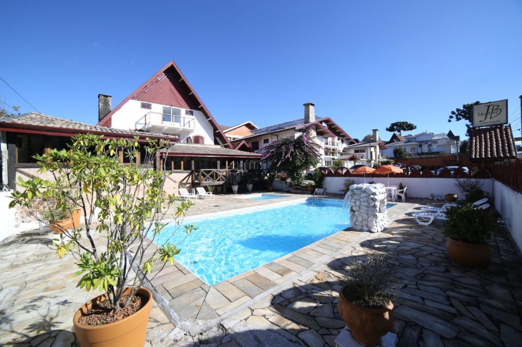 Piscina externa retangular do Hotel JB cercada por vasos de plantas e um pátio de pedra perto de um prédio com telhado vermelho em um dia ensolarado, para representar hotéis em Campos do Jordão.