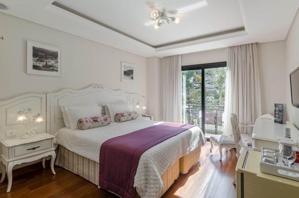Um quarto bem iluminado no Villa D'Biagy Premium com uma cama grande, móveis brancos, arte na parede, uma escrivaninha e uma janela com cortinas que deixam entrar a luz do sol.