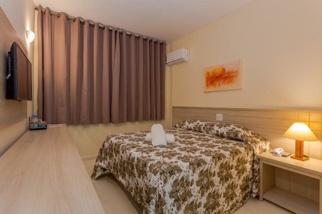 Quarto no Hotel Golden Park Campos com uma cama de casal, mesa de cabeceira com abajur, TV na parede, cortinas marrons e paredes bege.