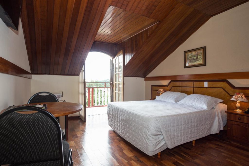 Quarto no Hotel Leão da Montanha com uma cama de casal, uma pequena mesa redonda, duas cadeiras e uma porta de sacada que permite a entrada de luz natural.