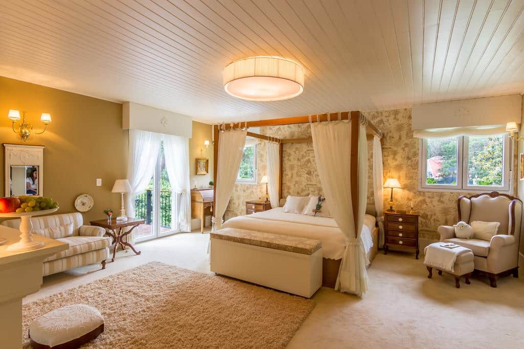 Quarto espaçoso na Casa Regaleira Hotel Boutique com cama de dossel, poltrona, sofá, escrivaninha, janelas grandes e iluminação aconchegante, para representar hotéis em Campos do Jordão.
