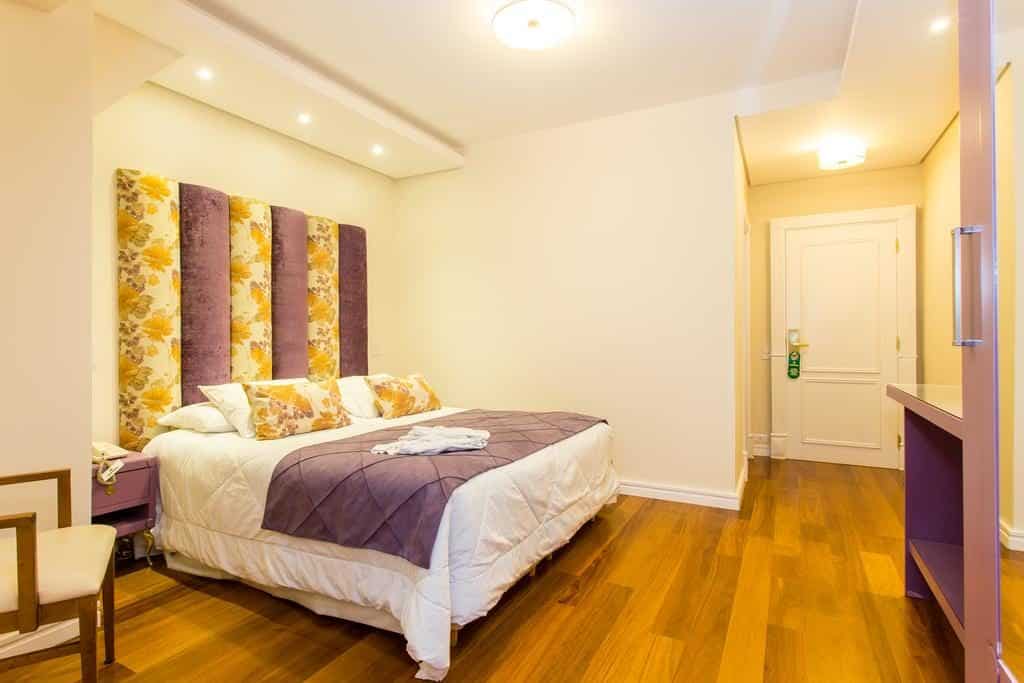 Um quarto no Hotel Serra da Estrela com uma cama de casal, cabeceira com estampa floral, piso de madeira e paredes brancas, com uma porta no final.