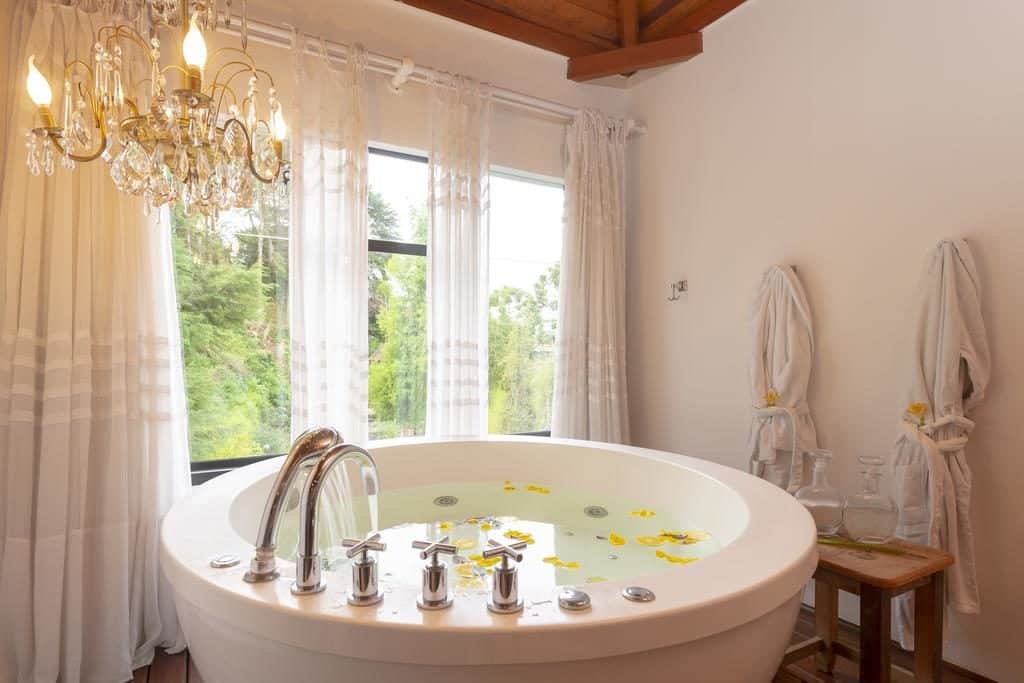 Uma banheira redonda branca cheia de água e pétalas de flores amarelas sob um lustre, perto de grandes janelas com cortinas transparentes, em um dos quartos no WE Hotel, para representar hotéis em Campos do Jordão.