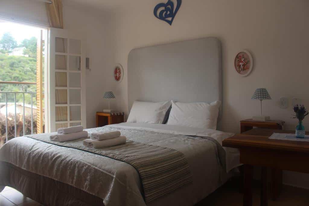 Uma cama bem arrumada com toalhas dobradas, mesas de cabeceira, luminárias e uma janela com vista para a vegetação em um quarto bem iluminado, em um dos quartos do Casarão da Ducha Hotel.