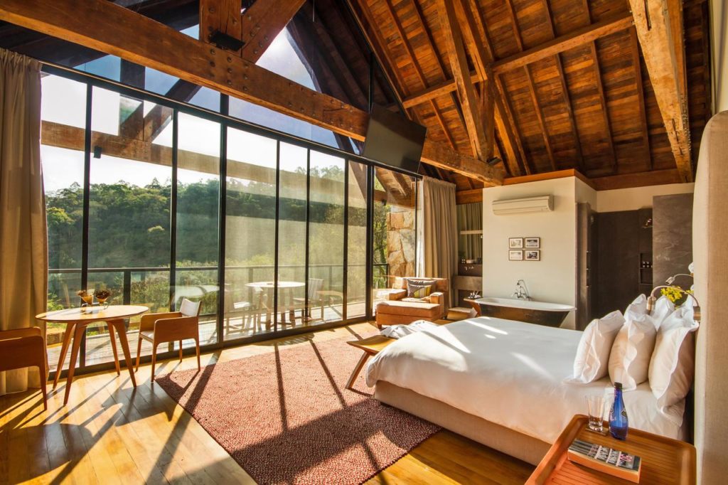 Um quarto iluminado pelo sol no Six Senses Botanique, com uma cama grande, área de estar, teto de madeira e portas de vidro que se abrem para uma varanda com vista panorâmica.