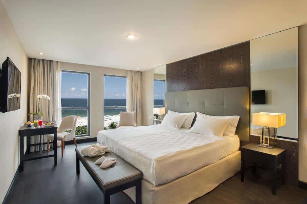 Quarto moderno no Windsor California Hotel com uma cama grande, escrivaninha, poltrona e duas janelas com vista para o mar e a praia, para representar hotéis em Copacabana.