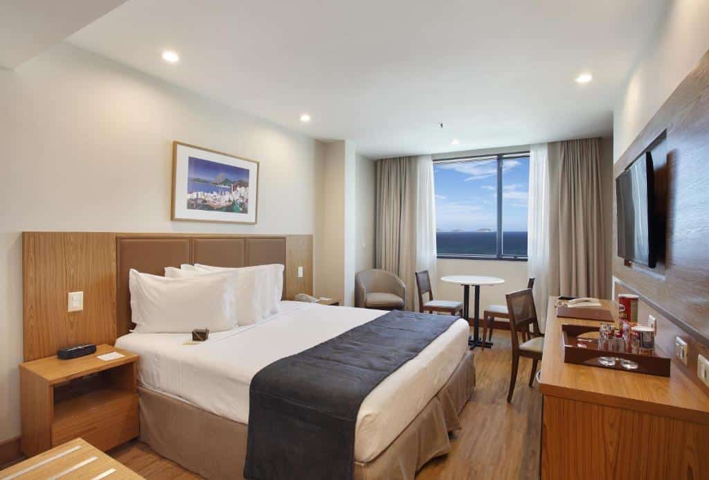 Quarto do Windsor Excelsior Copacabana com uma cama de casal, escrivaninha, TV, duas cadeiras, uma mesa e uma janela com vista para o mar, para representar hotéis em Copacabana.