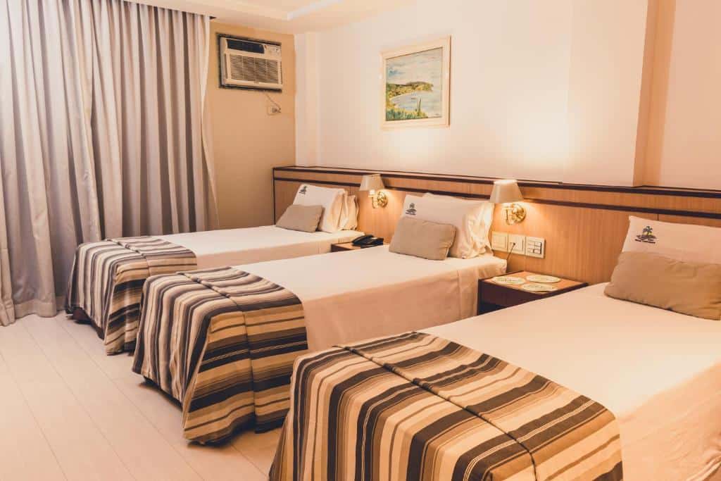 Um quarto do Riale Vilamar Copacabana com três camas de solteiro, roupa de cama listrada, luminárias de parede, um ar-condicionado e um quadro emoldurado, para representar hotéis em Copacabana.