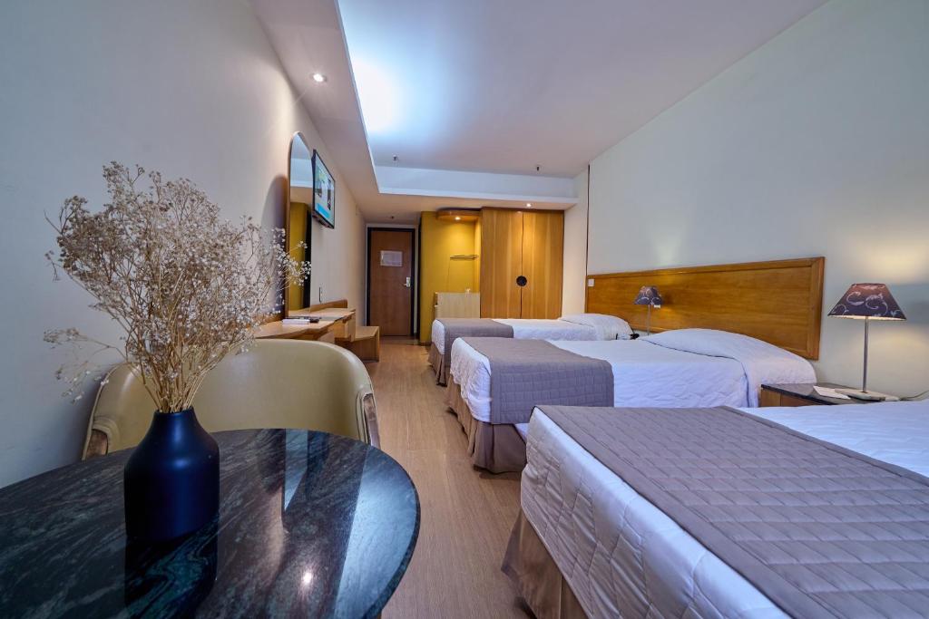 Quarto no South American Copacabana Hotel com três camas de solteiro, uma escrivaninha, TV e uma mesa redonda com um vaso de flores secas em primeiro plano, para representar hotéis em Copacabana.