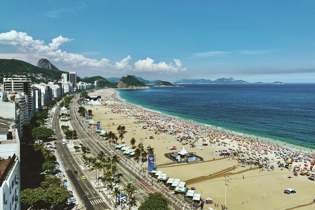 Vista aérea de uma praia movimentada próxima a uma cidade com prédios altos, montanhas e água azul clara sob um céu ensolarado, em Copacabana.