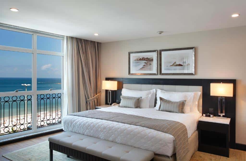 Quarto no Miramar Hotel by Windsor com uma cama grande, vista para o mar através de janelas do chão ao teto, decoração moderna e luminárias de cabeceira, para representar hotéis em Copacabana.