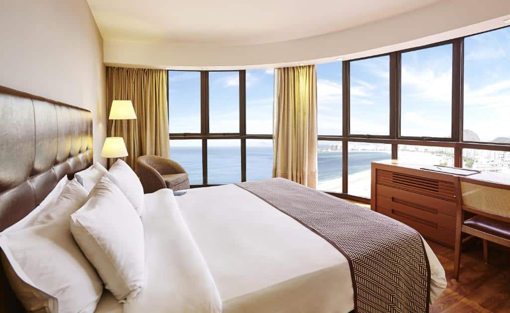 Quarto do PortoBay Rio de Janeiro com uma cama grande, poltrona, escrivaninha e janelas do chão ao teto com vista para o oceano e a cidade, para representar hotéis em Copacabana.