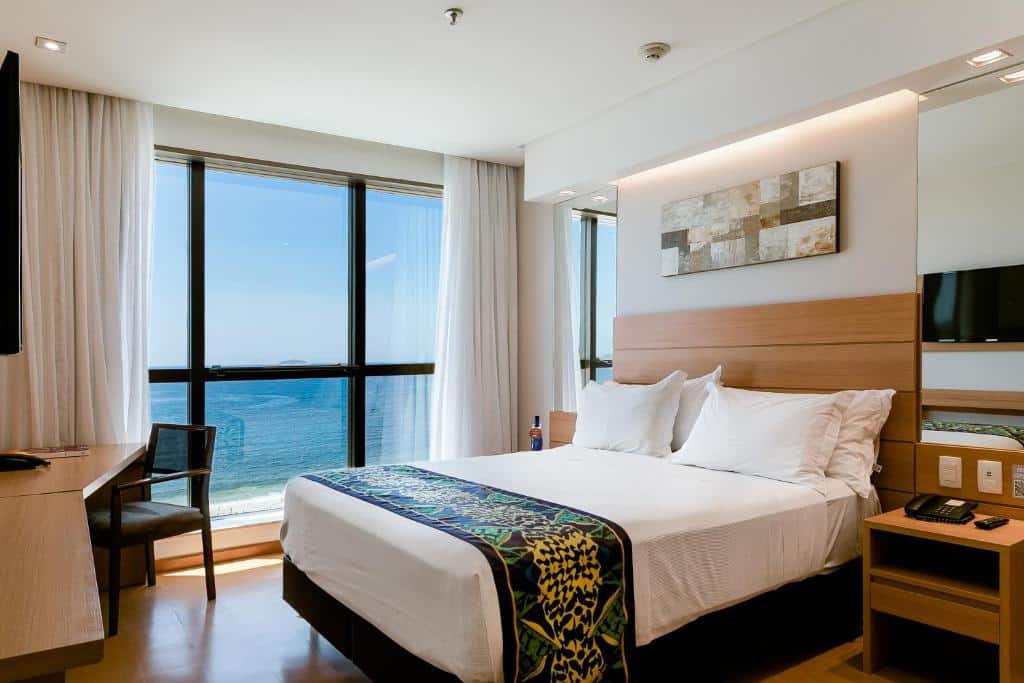 Um quarto do Arena Copacabana Hotel com uma cama de casal, escrivaninha e uma grande janela com vista para o mar sob a luz do dia, para representar hotéis em Copacabana.