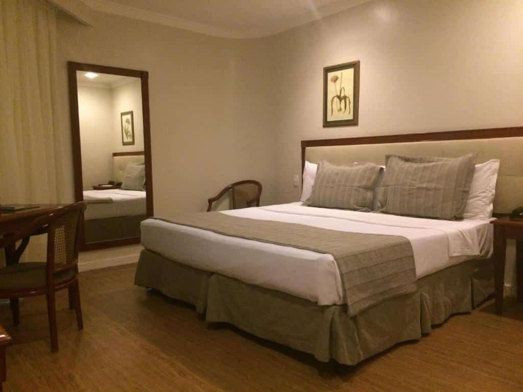 Uma cama bem arrumada com dois travesseiros e uma colcha bege em um quarto simples no Olinda Rio Hotel com um espelho e uma cadeira de madeira, para representar hotéis em Copacabana.