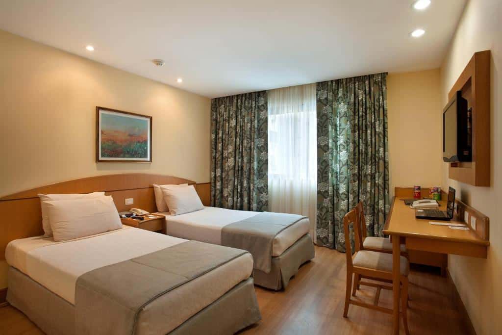 Quarto no Windsor Martinique com duas camas de solteiro, uma escrivaninha com cadeira, TV na parede, janela com cortinas e obras de arte emolduradas, para representar hotéis em Copacabana.