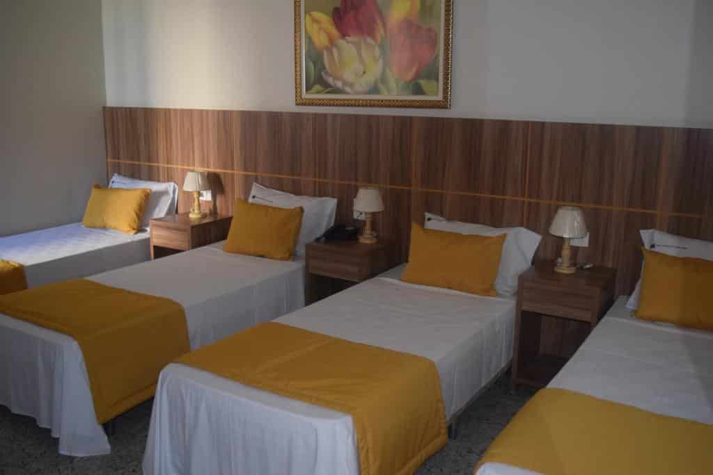 Quatro camas de solteiro com lençóis brancos e detalhes em amarelo estão dispostas em uma fileira em um quarto do Hotel Diplomata Copacabana com uma pintura floral acima, para representar hotéis em Copacabana.