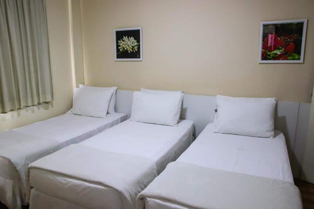 Três camas de solteiro com roupas de cama brancas dispostas lado a lado em um quarto do Hotel Acalanto com paredes claras e dois quadros emoldurados na parede.