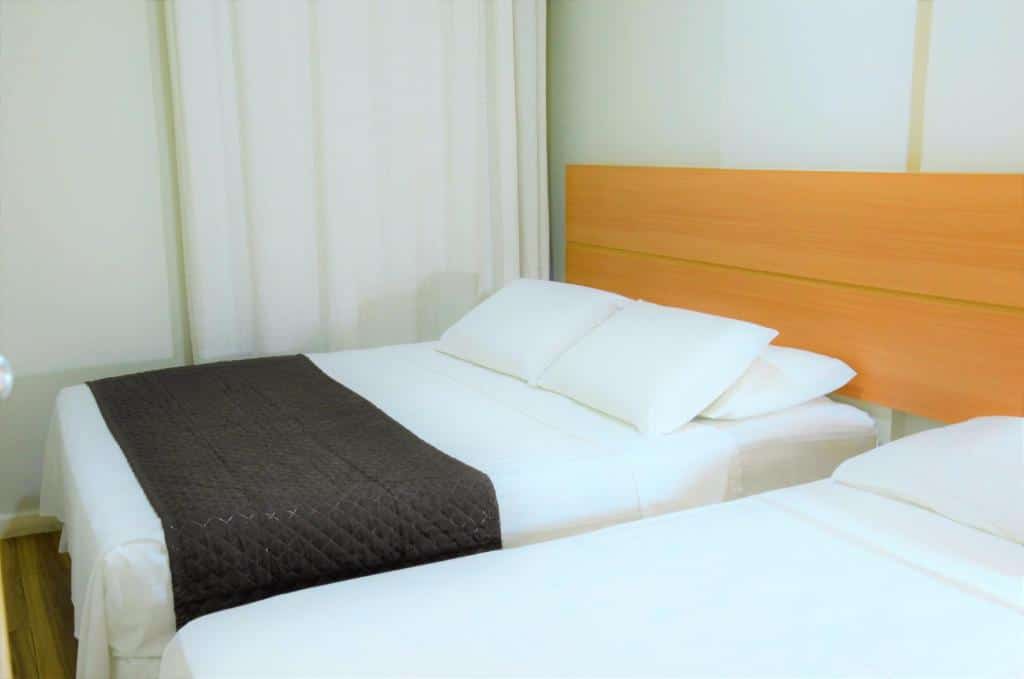Duas camas bem arrumadas com lençóis e travesseiros brancos; uma delas tem uma manta marrom acolchoada nos pés. Este é um dos quartos do Classe Apart Hotel em Feira de Santana.