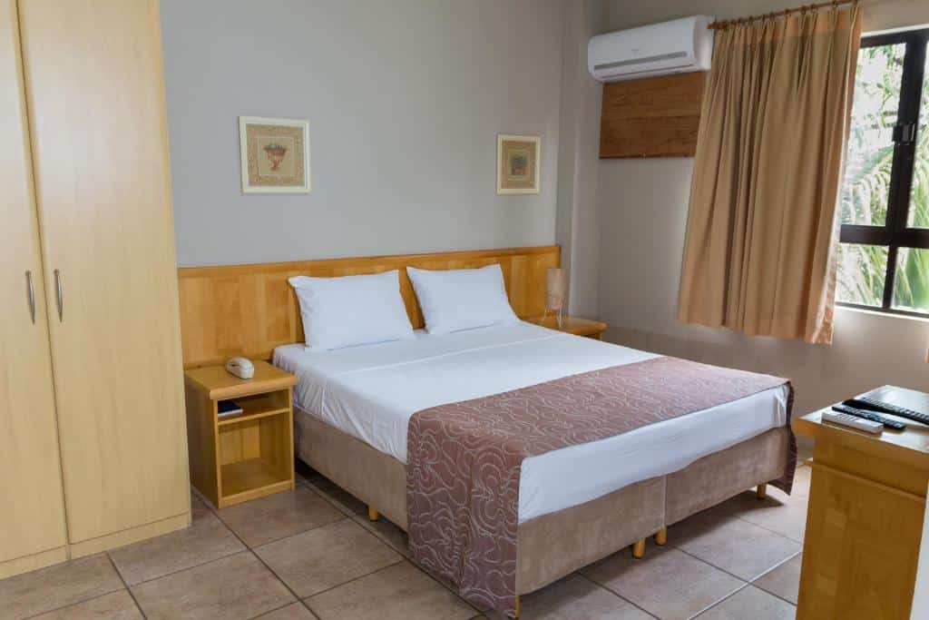 Um quarto arrumado do Maria Quitéria Hotel & Flat com uma cama de casal, móveis de madeira, piso de azulejo, janela com cortinas e ar-condicionado de parede.