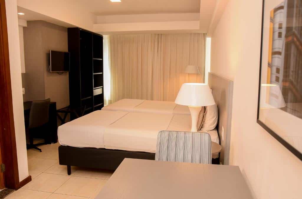 Um quarto do Executive Hotel com duas camas de solteiro, uma escrivaninha, cadeira, TV, abajur e cortinas cobrindo uma grande janela.