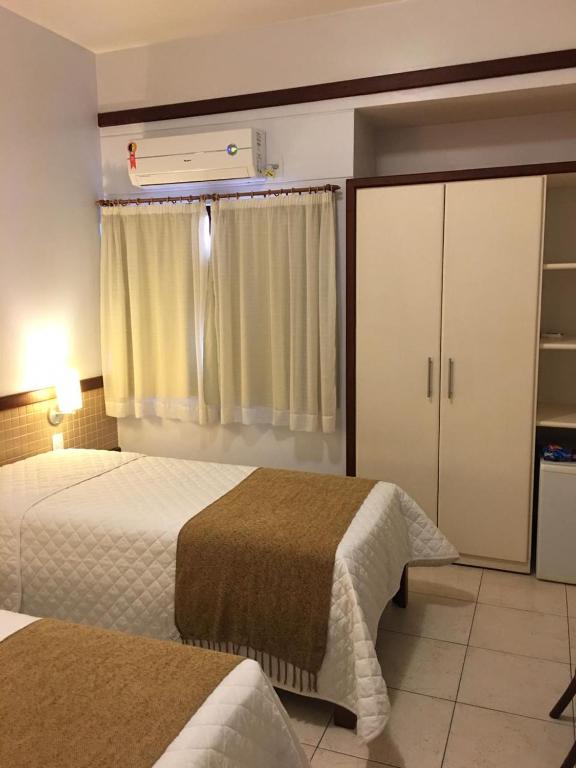 Um quarto de hotel com duas camas de solteiro, cobertores bege, um guarda-roupa, piso de azulejo e uma janela com cortinas fechadas.