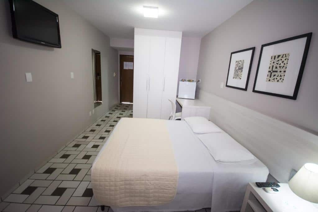 Uma cama bem arrumada em um quarto simples do Hotel São Domingos com móveis brancos, piso de azulejo, TV, guarda-roupa e arte na parede.