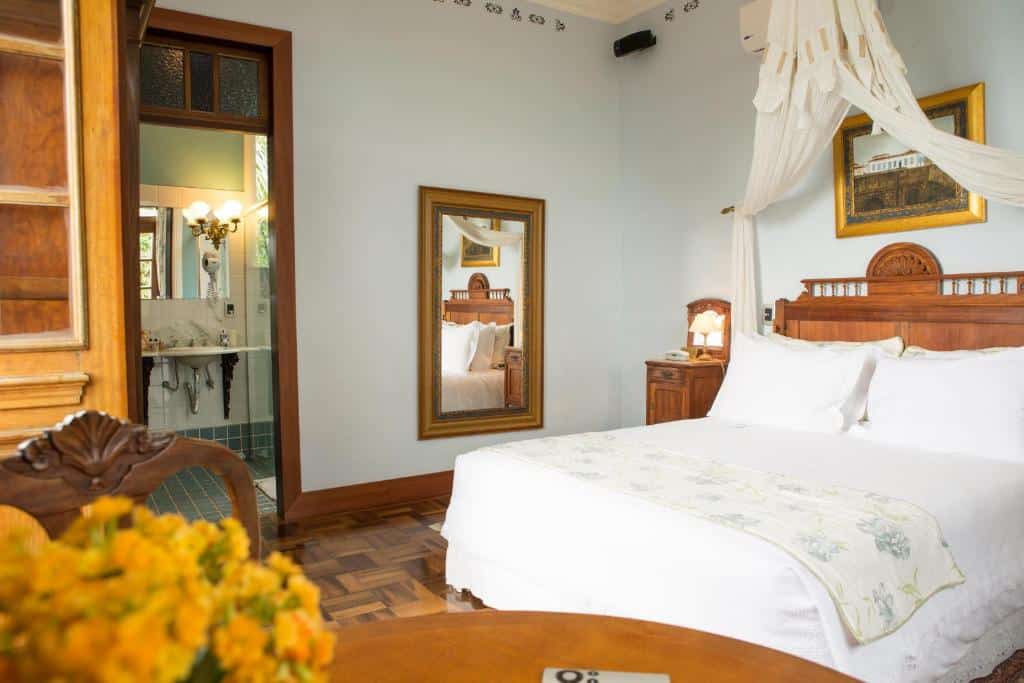 Uma cama bem arrumada em um quarto aconchegante com móveis de madeira, um espelho e vista para um banheiro com uma arandela de parede iluminada.. Um dos hotéis em Florianópolis.