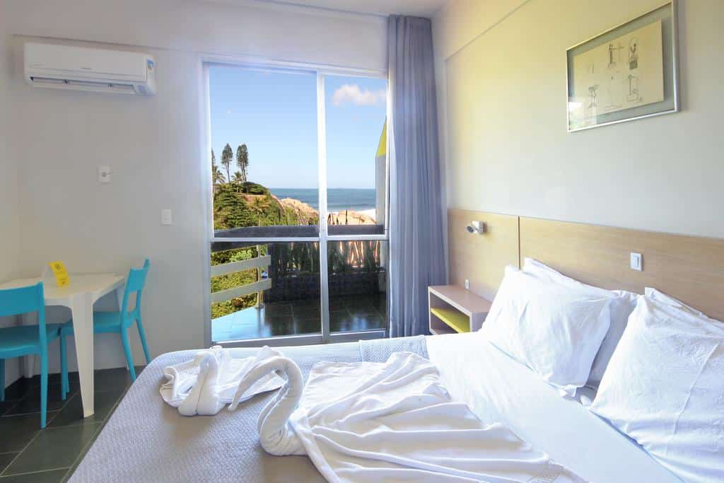 Quarto de hotel bem iluminado com uma cama feita, toalha de cisne, mesa pequena com duas cadeiras e uma janela com vista para o mar. Um dos hotéis em Florianópolis.