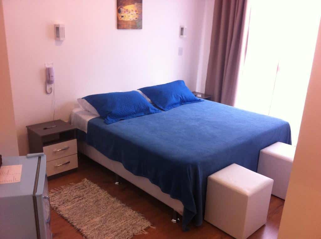 Uma cama bem arrumada com roupa de cama azul, dois travesseiros azuis, dois pufes brancos e duas mesas de cabeceira em um quarto bem iluminado. Um dos hotéis em Florianópolis.