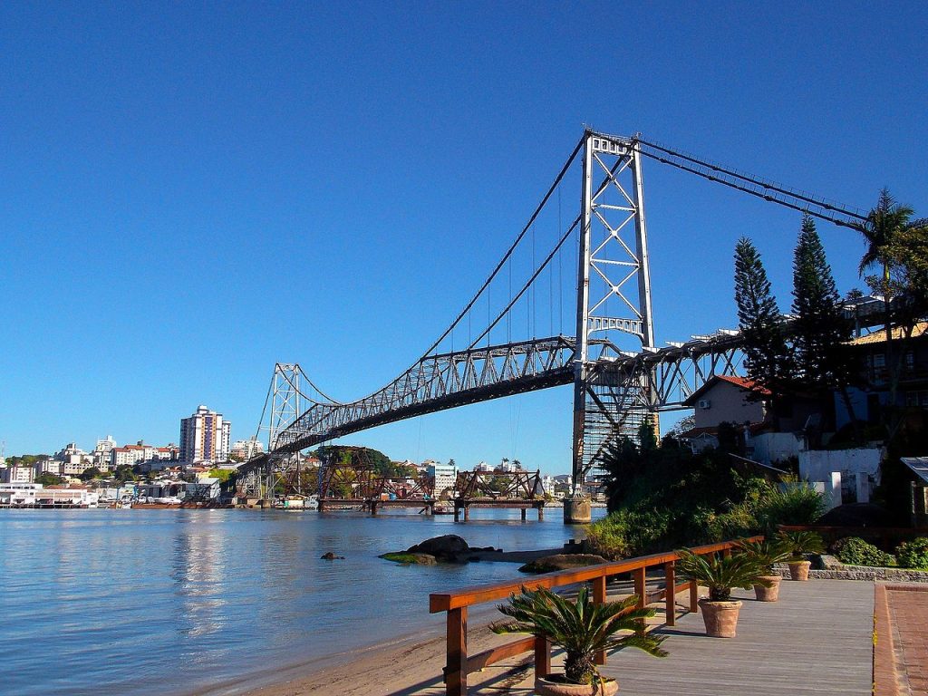 Uma grande ponte suspensa se estende sobre um corpo de água com uma paisagem urbana e um céu azul claro ao fundo.