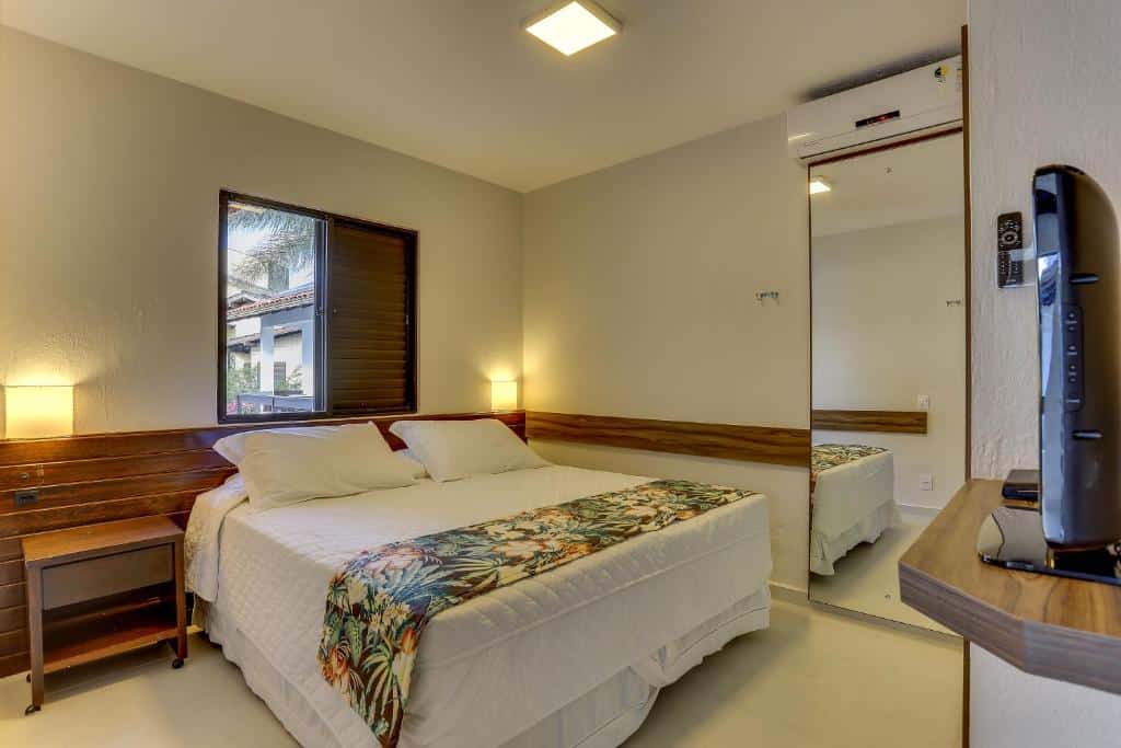 Um quarto de hotel com uma cama de casal, duas mesinhas de cabeceira, um espelho de parede, uma janela e uma TV em uma prateleira de canto.