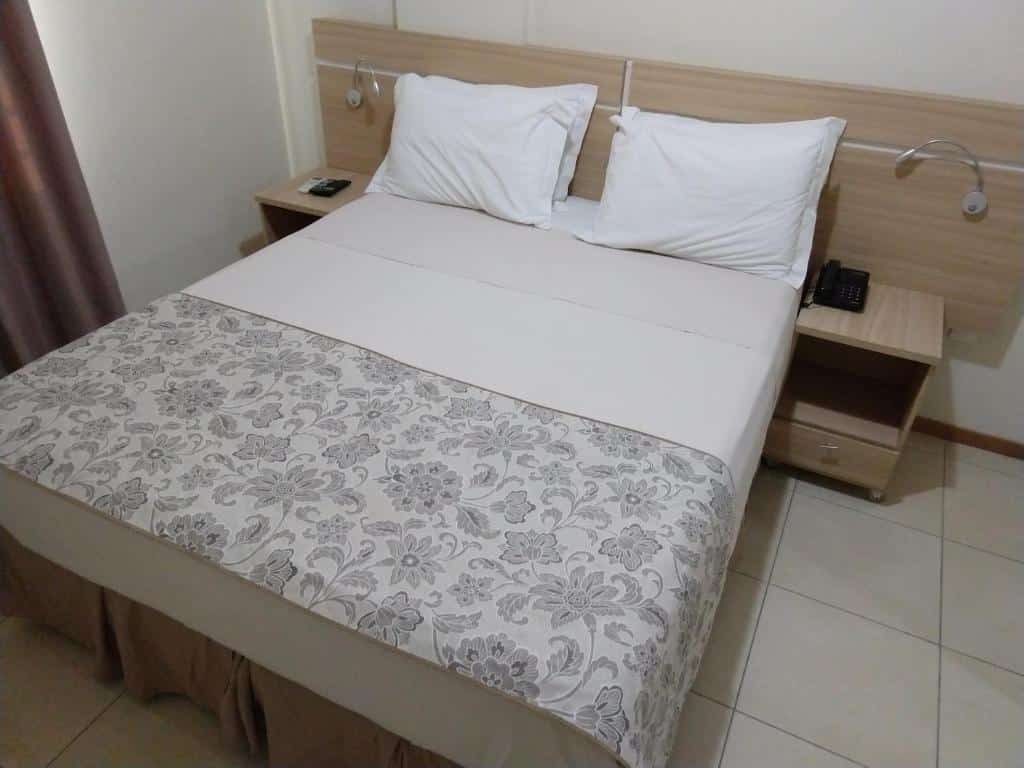 Uma cama bem arrumada com dois travesseiros, cobertor com estampa floral e mesinhas de cabeceira de cada lado, em um quarto de azulejos. Este é um dos hotéis em Florianópolis.