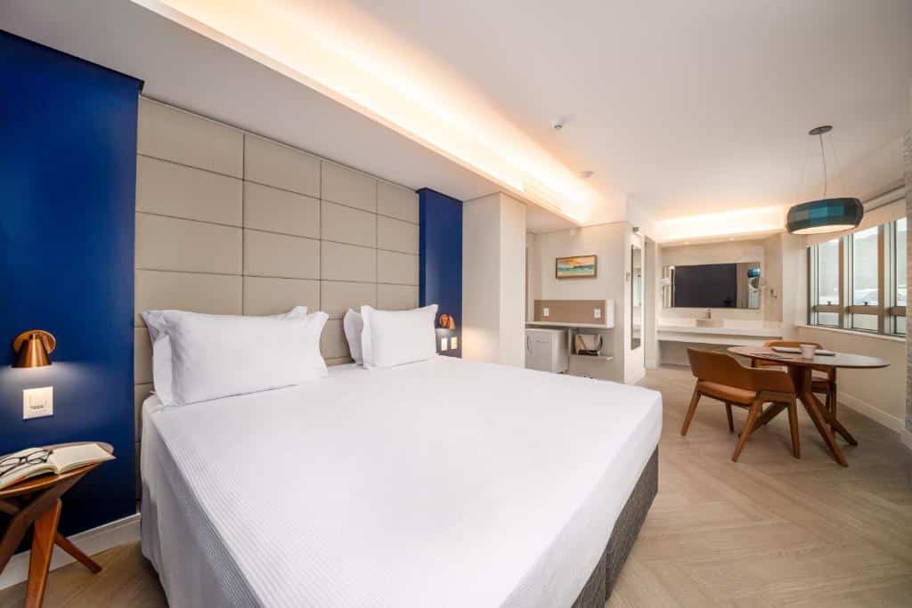 Quarto de hotel moderno com uma cama grande, paredes com detalhes em azul, área de trabalho, mesa de jantar e uma TV de tela plana. Um dos hotéis em Florianópolis.