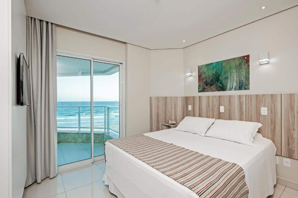 Um quarto de hotel com uma cama de casal, arte na parede, TV e uma varanda com vista para o oceano. Um dos hotéis em Florianópolis.