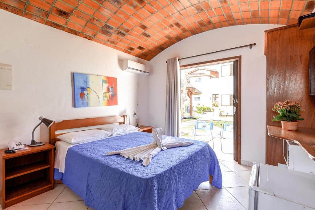 Um quarto de hotel com colcha azul, arte em toalha na cama, escrivaninha, móveis de madeira e uma porta que dá para o exterior. Um dos hotéis em Florianópolis.