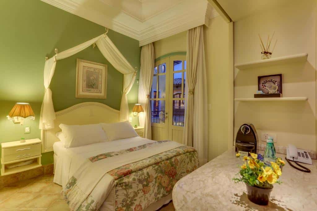 Um quarto aconchegante com cama de casal, roupa de cama floral, parede com detalhes em verde, escrivaninha e portas francesas que dão para uma varanda. Um dos hotéis em Florianópolis.