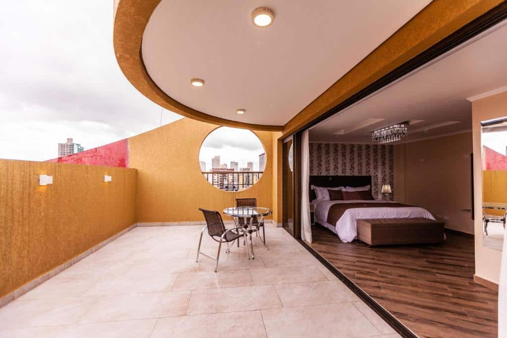 Quarto do Foz Plaza Hotel que mostra do lado direito o quarto com a cama de casal e um baú nos pés. Já do lado esquerdo tem a varanda do quarto com uma mesa redonda e duas cadeiras.