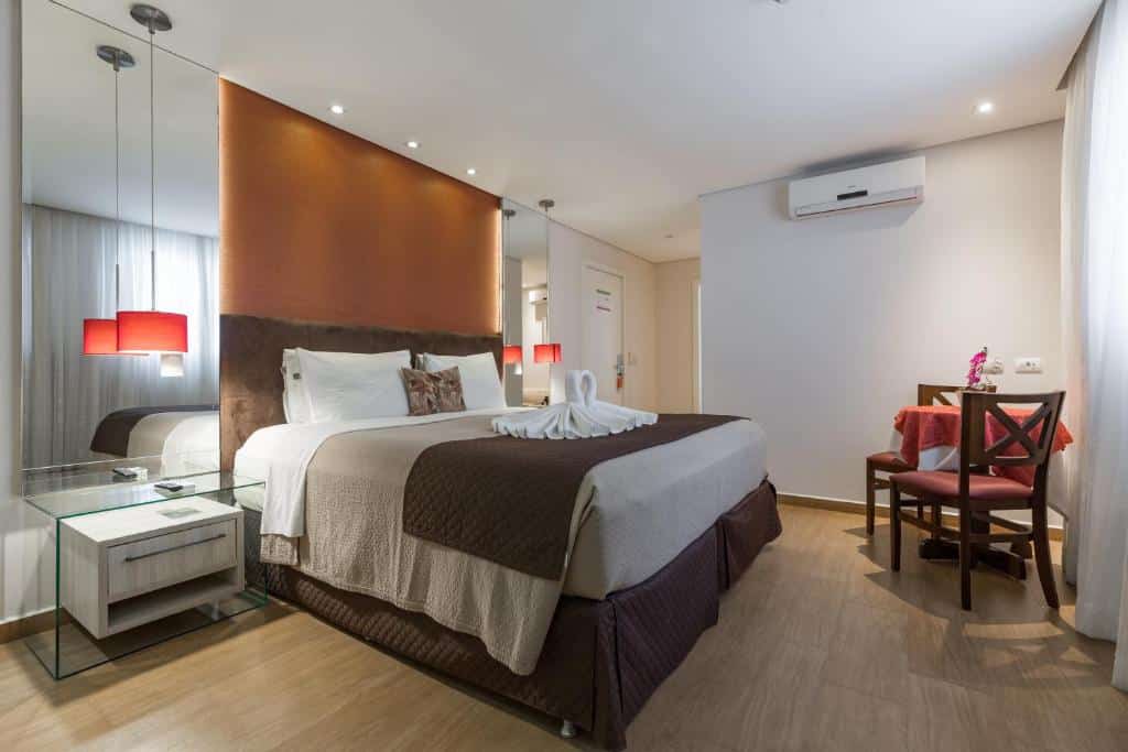 Quarto do Tarobá Hotel com uma cama de casal box e do lado direito uma mesa de cabeceira de vidro. Na frente da cama tem uma mesa com duas cadeiras e uma cortina. Já na parede esquerda tem um ventilador e um corredor com uma porta.