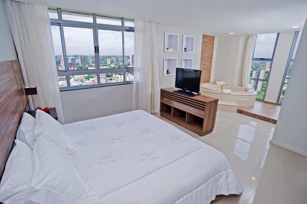 Quarto do Wyndham Golden Foz Suítes com uma cama de casal com uma tv em frente e do lado esquerdo tem uma janela de vidro com cortinas. Atrás da tv tem uma banheira e outras janelas de vidro.