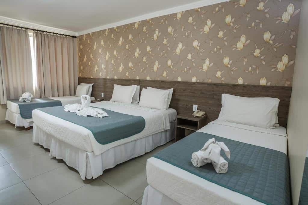 Quarto do Cataratas Park Hotel com uma cama de casal no centro e duas camas de solteiro uma de cada lado da cama de casal. Separando as camas tem duas mesas de cabeceira e do lado da cama de solteiro do fundo tem uma janela com cortinas.